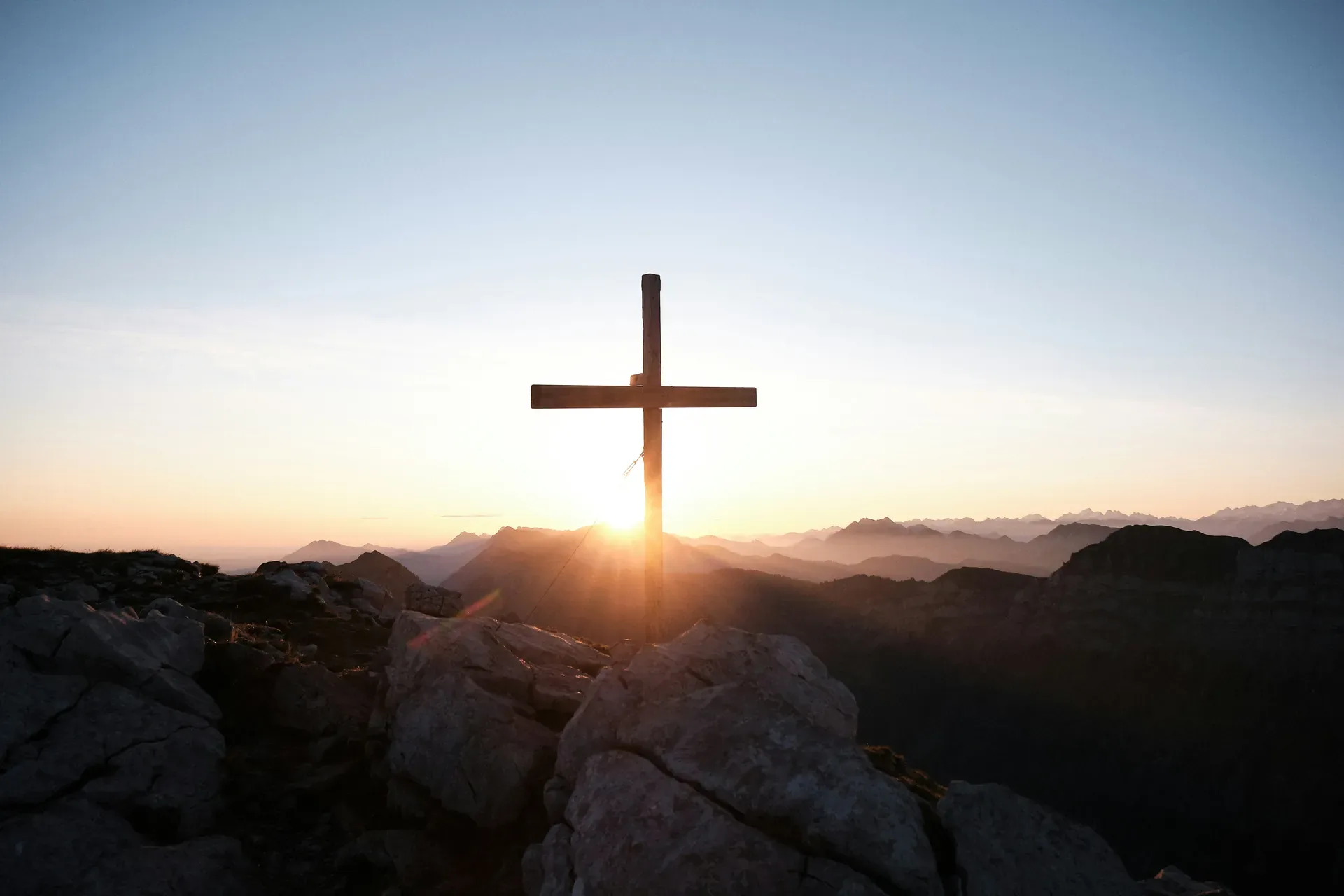 Wooden cross on a rocky mountain summit at sunrise with mountain peaks in the distance