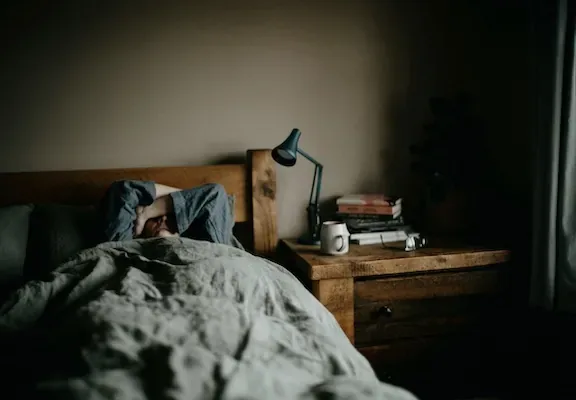 Person resting in bed beside a wooden nightstand with a lamp, books, and a mug in a quiet room